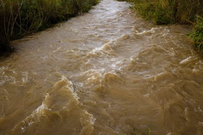 Rio barrento correndo com força por uma paisagem natural após chuvas intensas, com sinais de erosão nas margens